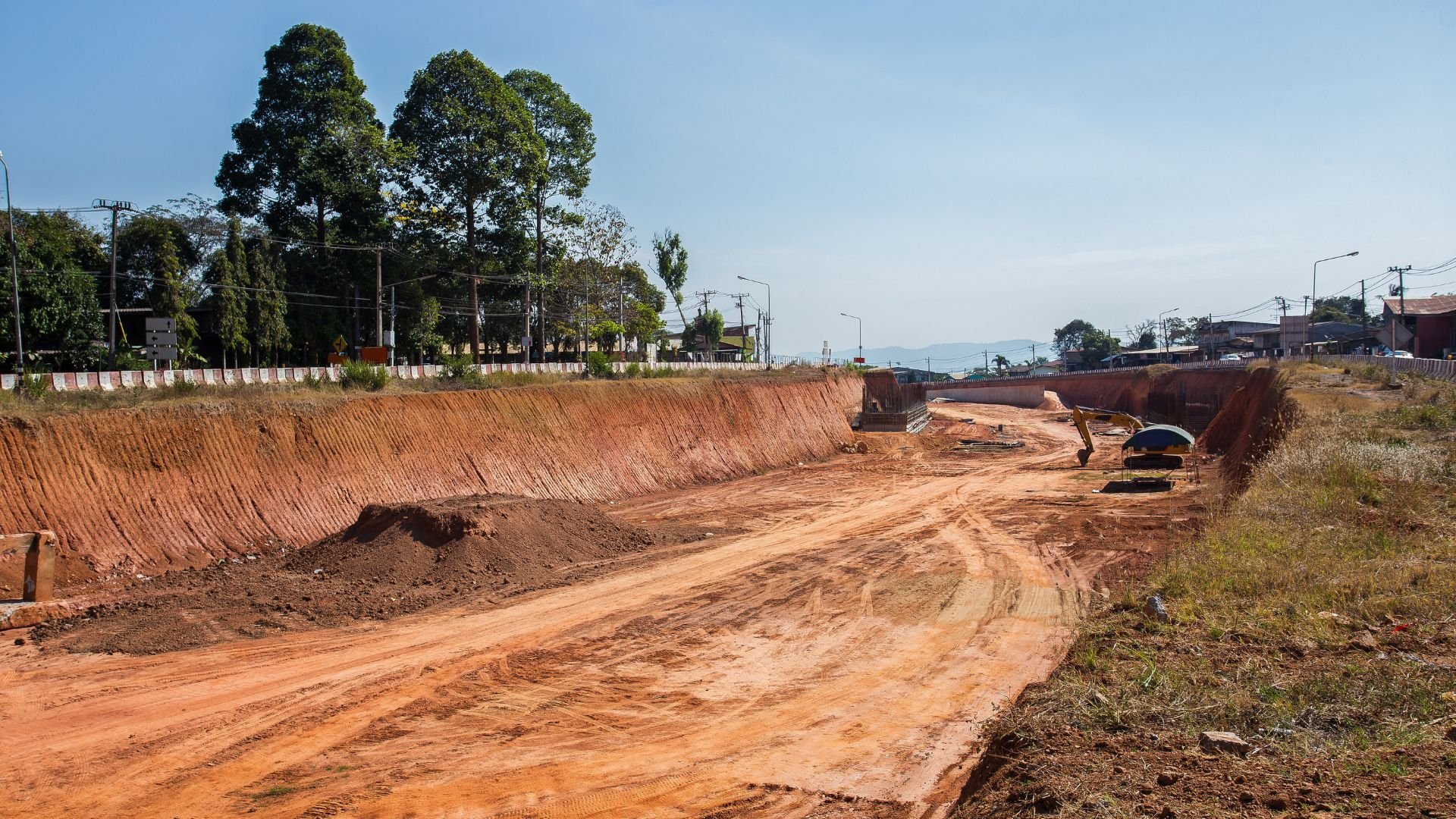 Road construction site with red earth, excavator, and trees in background